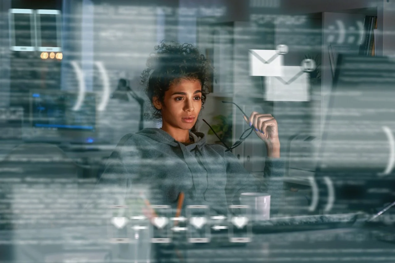A young woman with curly dark hair sitting at a desk in an office, viewed through a glass window with reflections of the office environment and icons of an email and chat message.