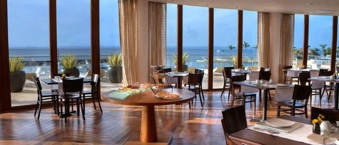 Indoor restaurant with large windows overlooking the beach, tables and chairs set for dining.