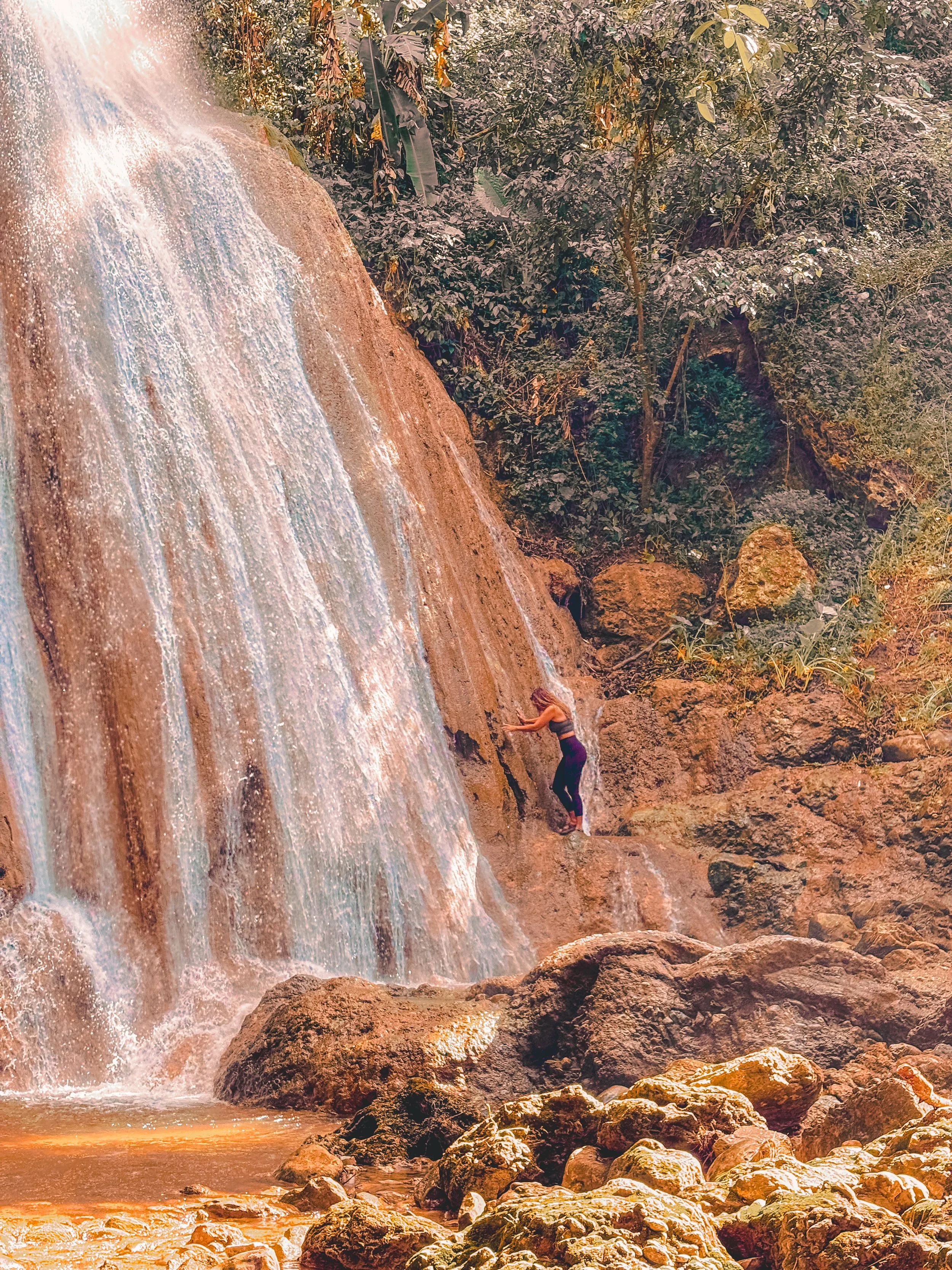 A woman climbing on rocks near a waterfall in a lush, green forest.