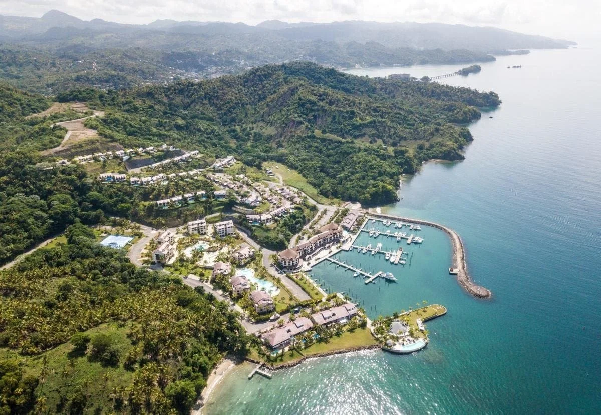 Aerial view of a coastal resort with a marina, surrounding lush green hills, residential buildings, and a beach along the shoreline.