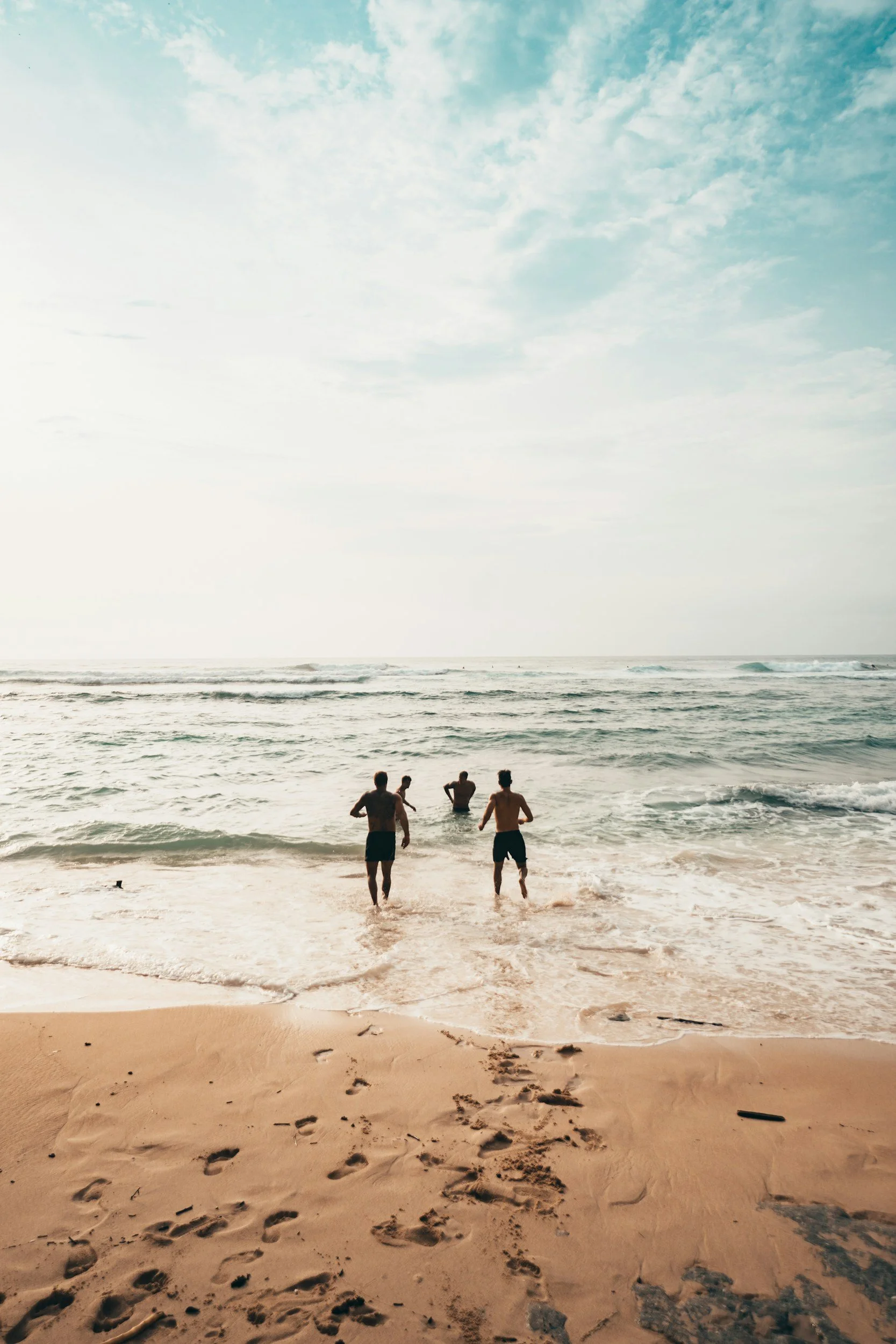 Four people running into the ocean at a beach, footprints in the sand, cloudy sky.