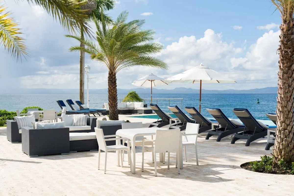 Beachside seating area with white and black outdoor furniture, palm trees, umbrellas, and lounge chairs overlooking the ocean, with mountains in the distance, under a partly cloudy sky.