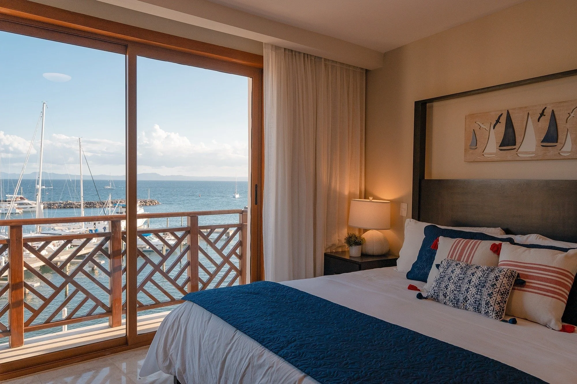 Bedroom with large sliding glass door overlooking a marina with sailboats and the ocean, featuring a bed with white and blue bedding, a lamp, and nautical decor.