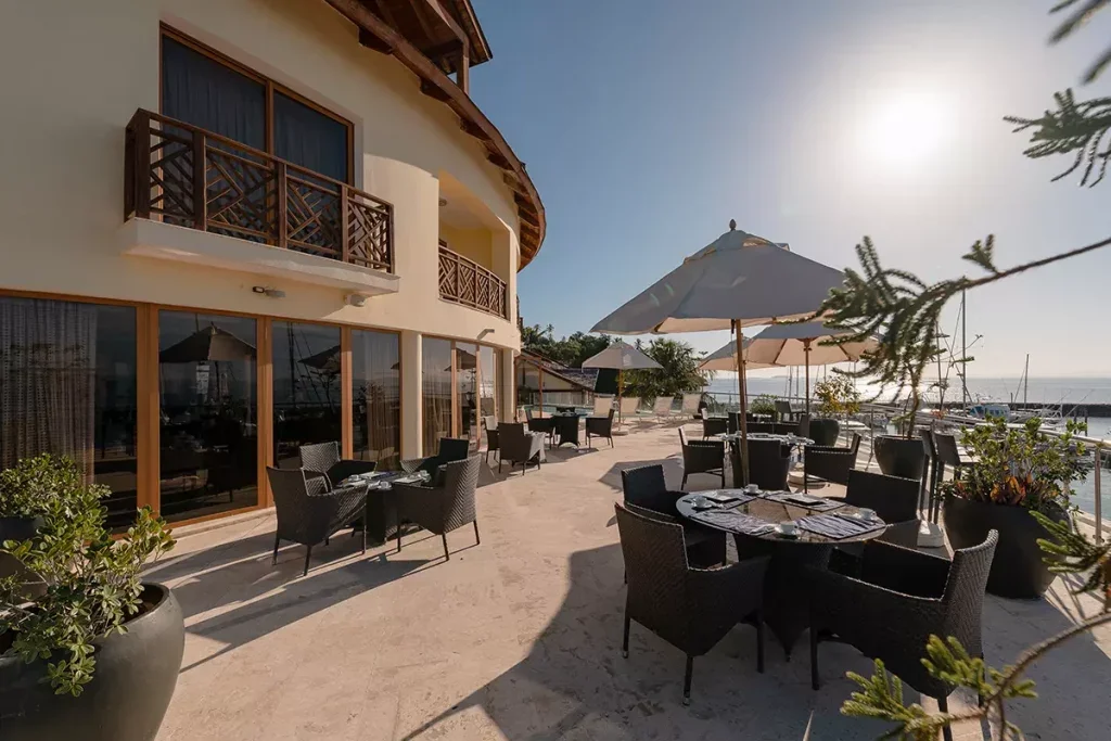 Outdoor restaurant patio with tables, chairs, and large umbrellas overlooking the water during daytime.