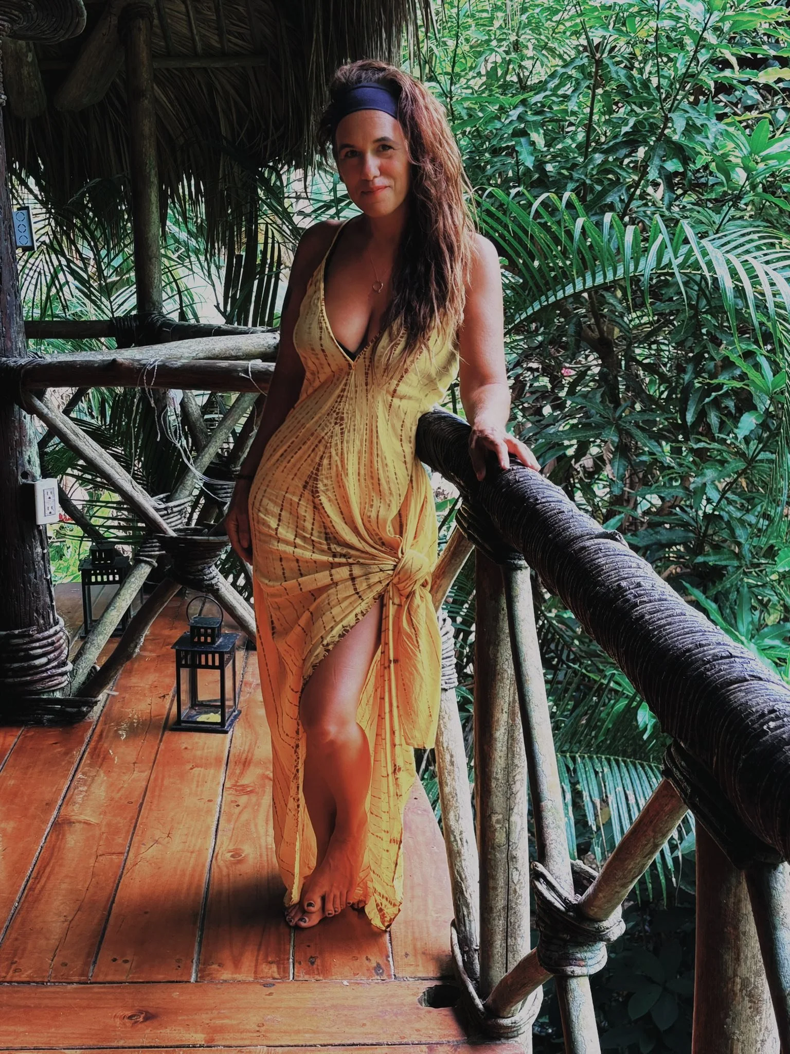 Woman on wooden balcony with greenery in the background, wearing a yellow dress and headband.