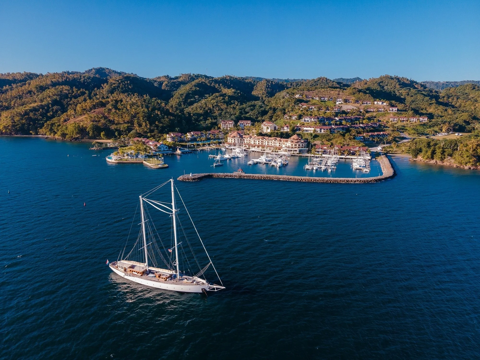 An aerial view of a marina with boats docked, surrounded by lush green hills and residential buildings under a clear blue sky.