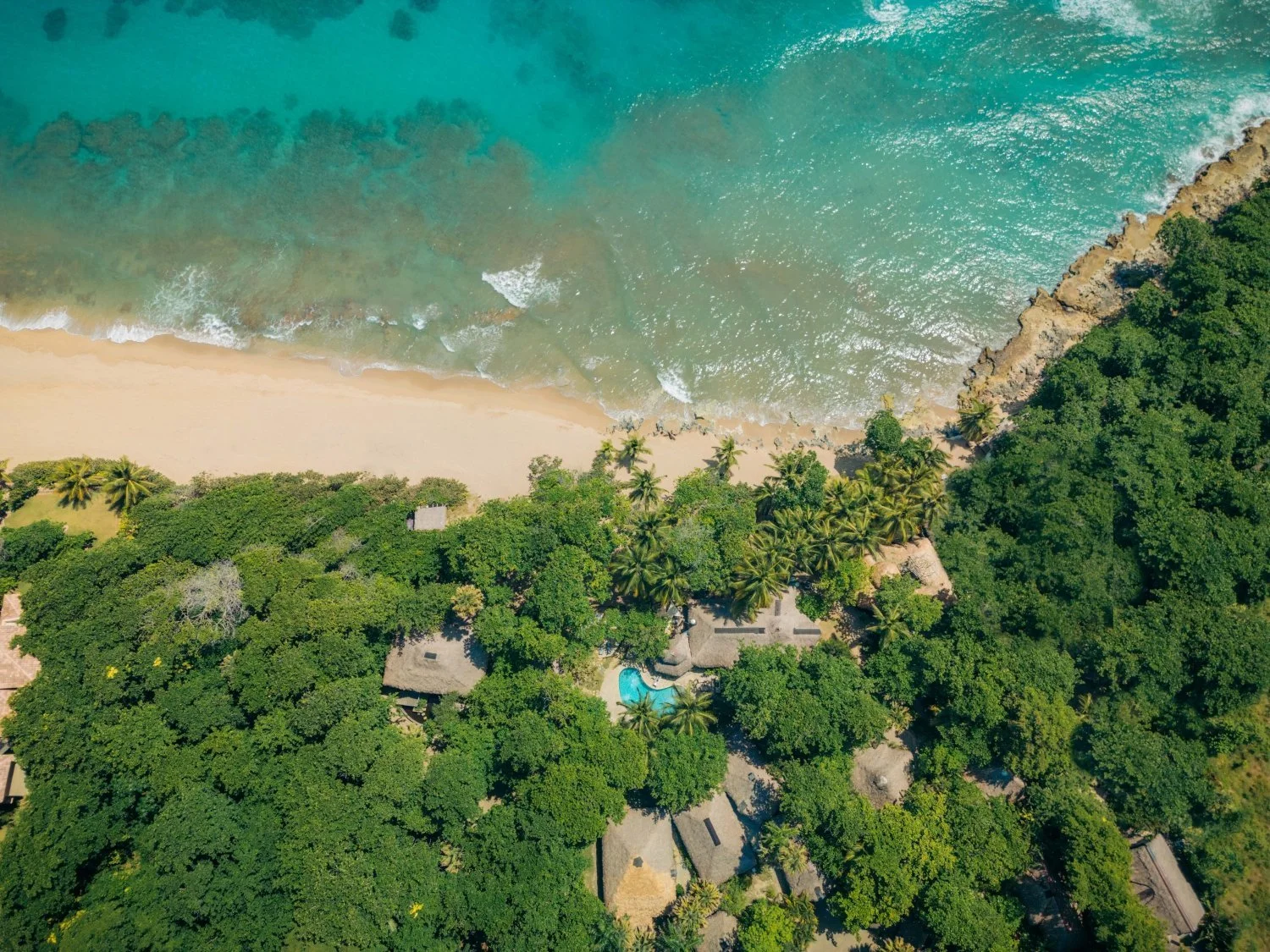 Aerial view of a tropical beach with turquoise water, sandy shoreline, lush green trees, and thatched-roof structures.