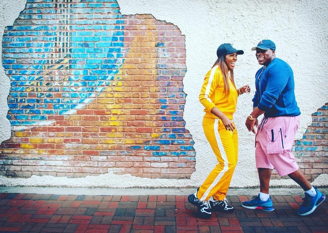 Two people, a woman in a yellow tracksuit and a man in a blue hoodie and pink shorts, standing and laughing in front of a brick wall with colorful graffiti.