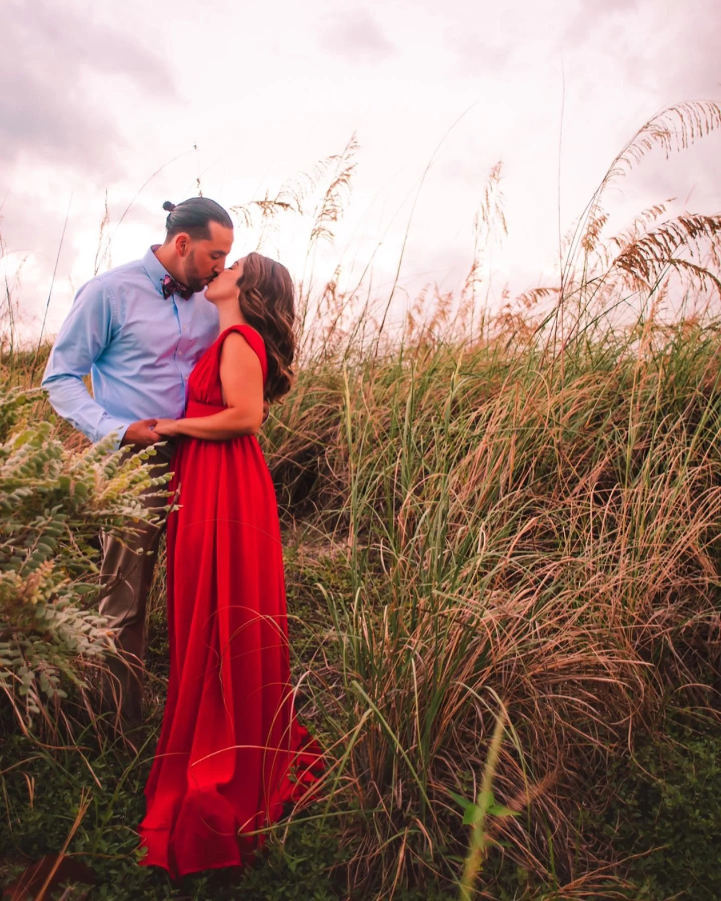 A couple sharing a kiss in a field of tall grass, with the woman wearing a long red dress and the man in a light blue shirt and bow tie.