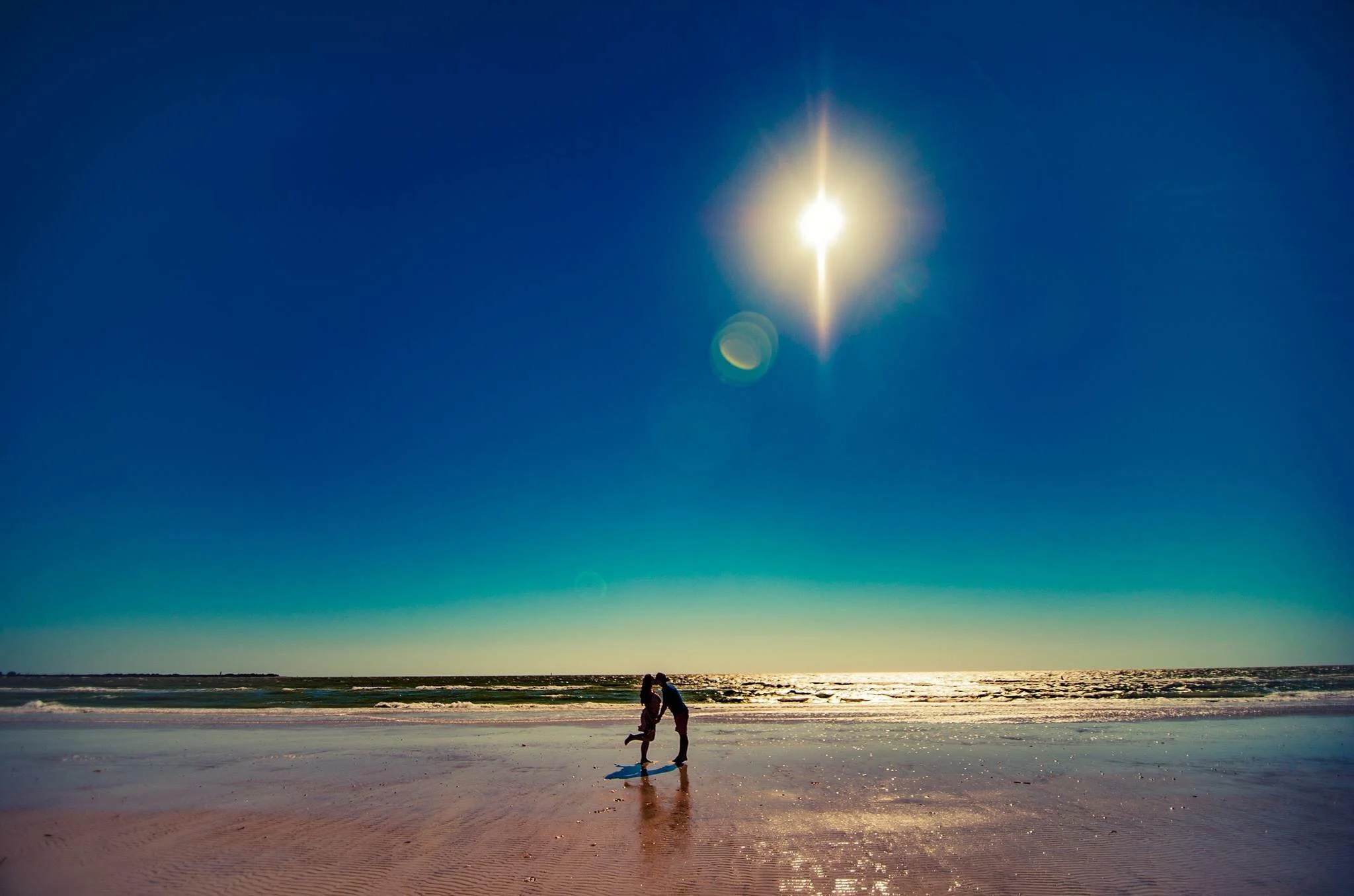 Couple kissing on the beach with the ocean and bright sun in the sky.