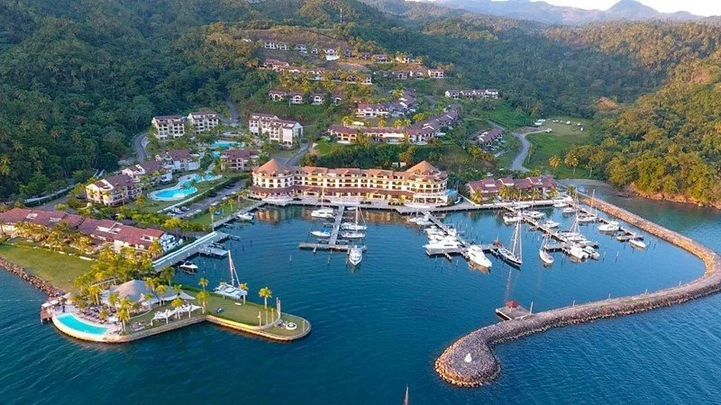 Aerial view of a marina with yachts, a resort with a pool, and residential buildings surrounded by green hills