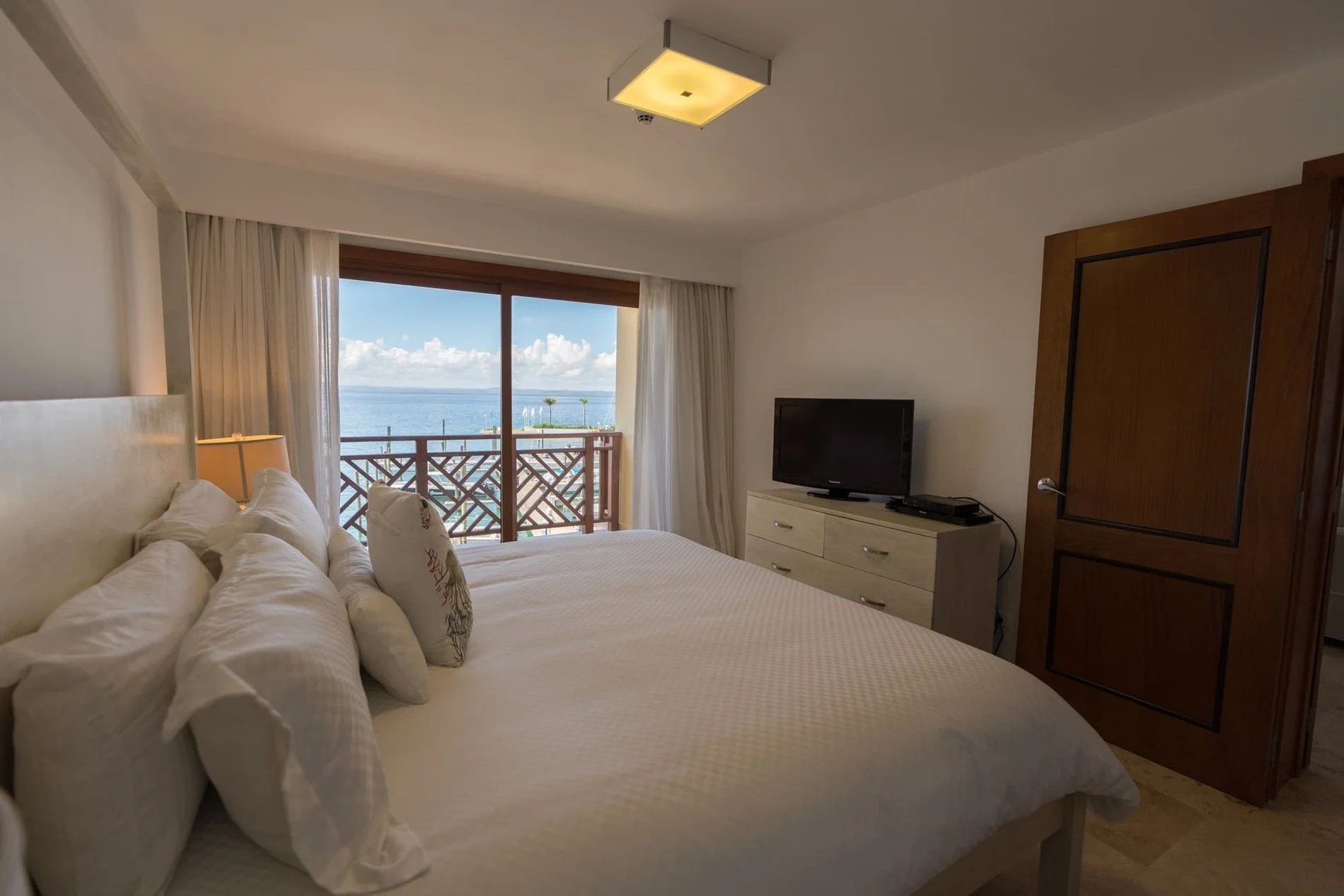 Hotel room with a large bed, a window with a view of the ocean and a balcony, a television on a dresser, and a wooden door.