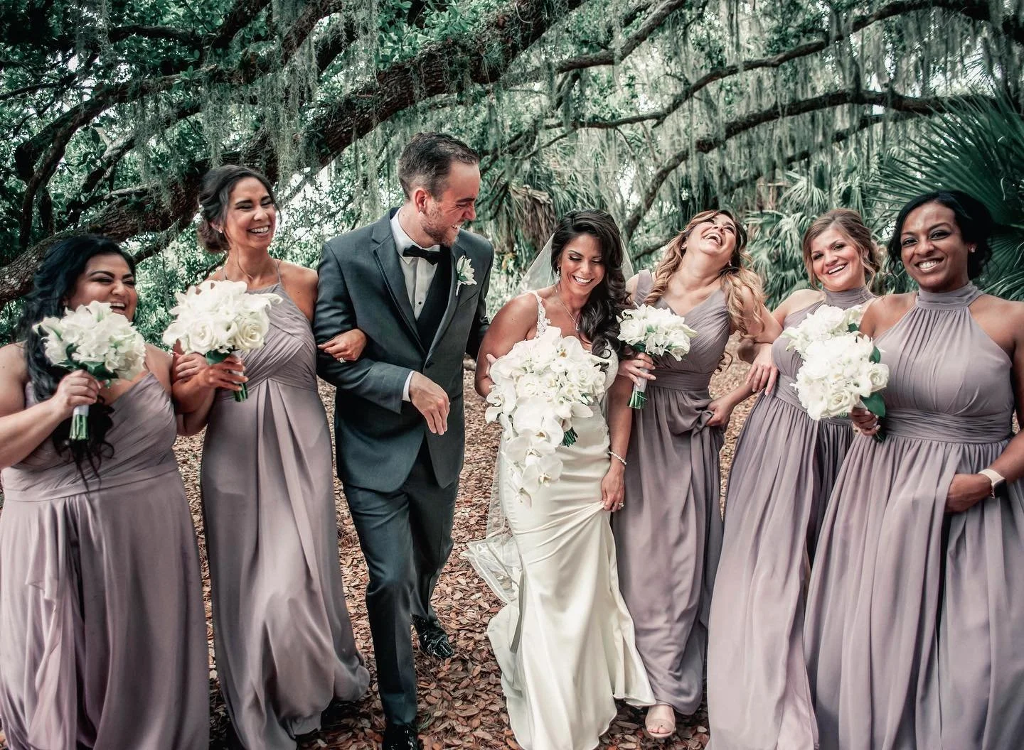 Wedding party standing outdoors under trees, smiling and laughing, with the bride and groom in the center holding bouquets of white flowers, surrounded by bridesmaids in matching taupe dresses also holding bouquets.