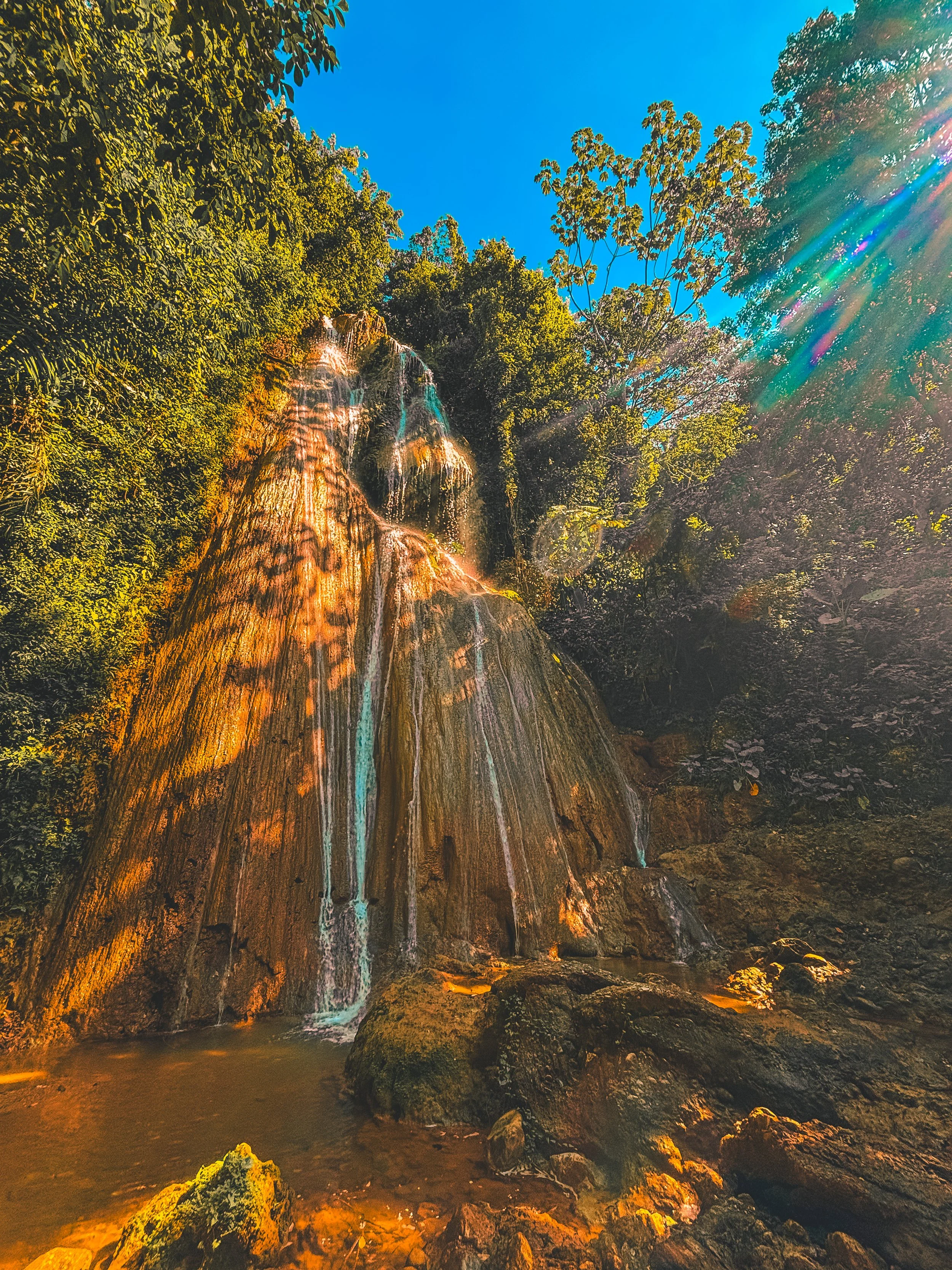A waterfall flowing down a rocky cliff surrounded by lush green trees, with sunlight creating a rainbow-like effect in the top right corner under a clear blue sky.