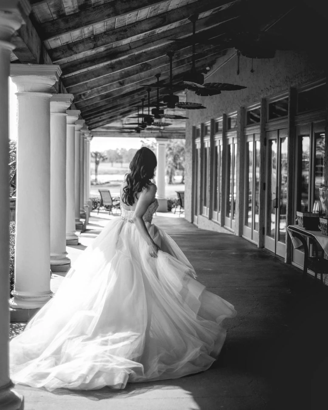 A woman in a wedding dress walking on a covered porch with columns and large glass doors, with outdoor patio furniture and trees visible outside.