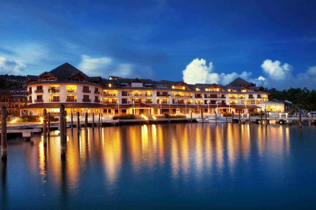 Night view of a waterfront hotel with multiple floors, balconies, and warm indoor lighting, reflecting on the calm water and docked boats in the foreground.