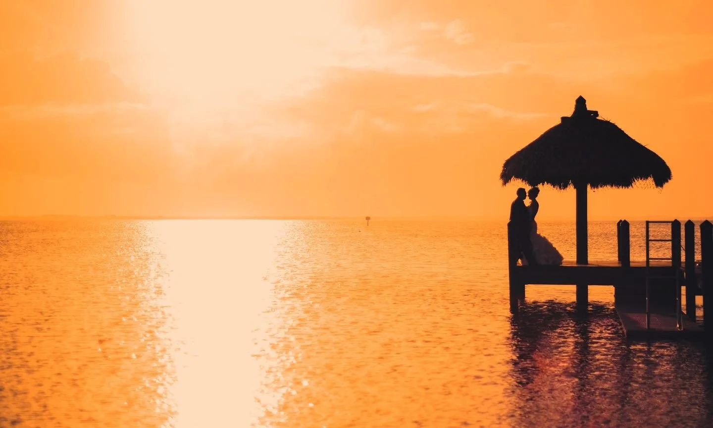 Silhouettes of a couple embracing under a thatched umbrella on a dock at sunset over a calm body of water.