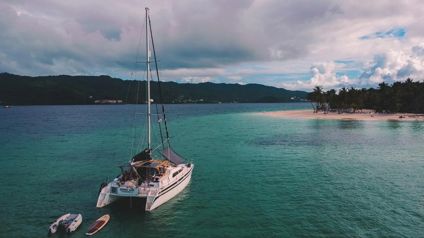 A sailboat anchored near a tropical island with palm trees and a sandy beach, calm turquoise water, and a cloudy sky with patches of blue in the background.