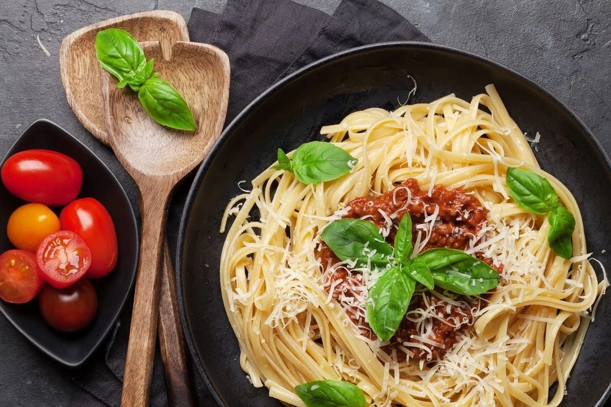 A plate of spaghetti with tomato sauce, shredded cheese, and fresh basil leaves, alongside a small black dish of cherry tomatoes and a wooden spoon with basil leaves on a dark surface.