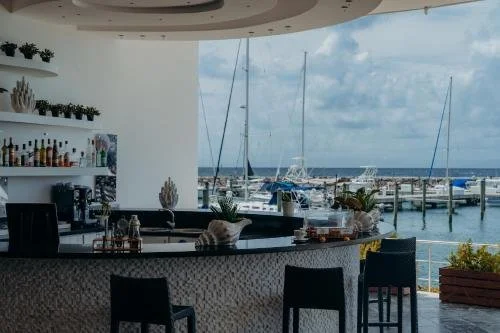 View of a marina with sailboats docked, seen from an indoor bar area with a curved counter, bar stools, and potted plants.