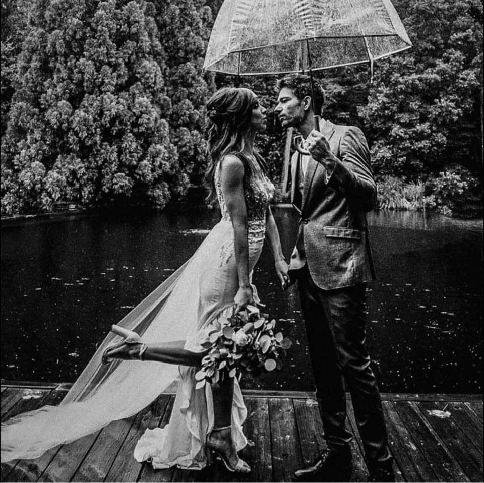 A bride and groom stand close together under a transparent umbrella on a wooden dock beside a lake, with trees in the background, during a rainy day.