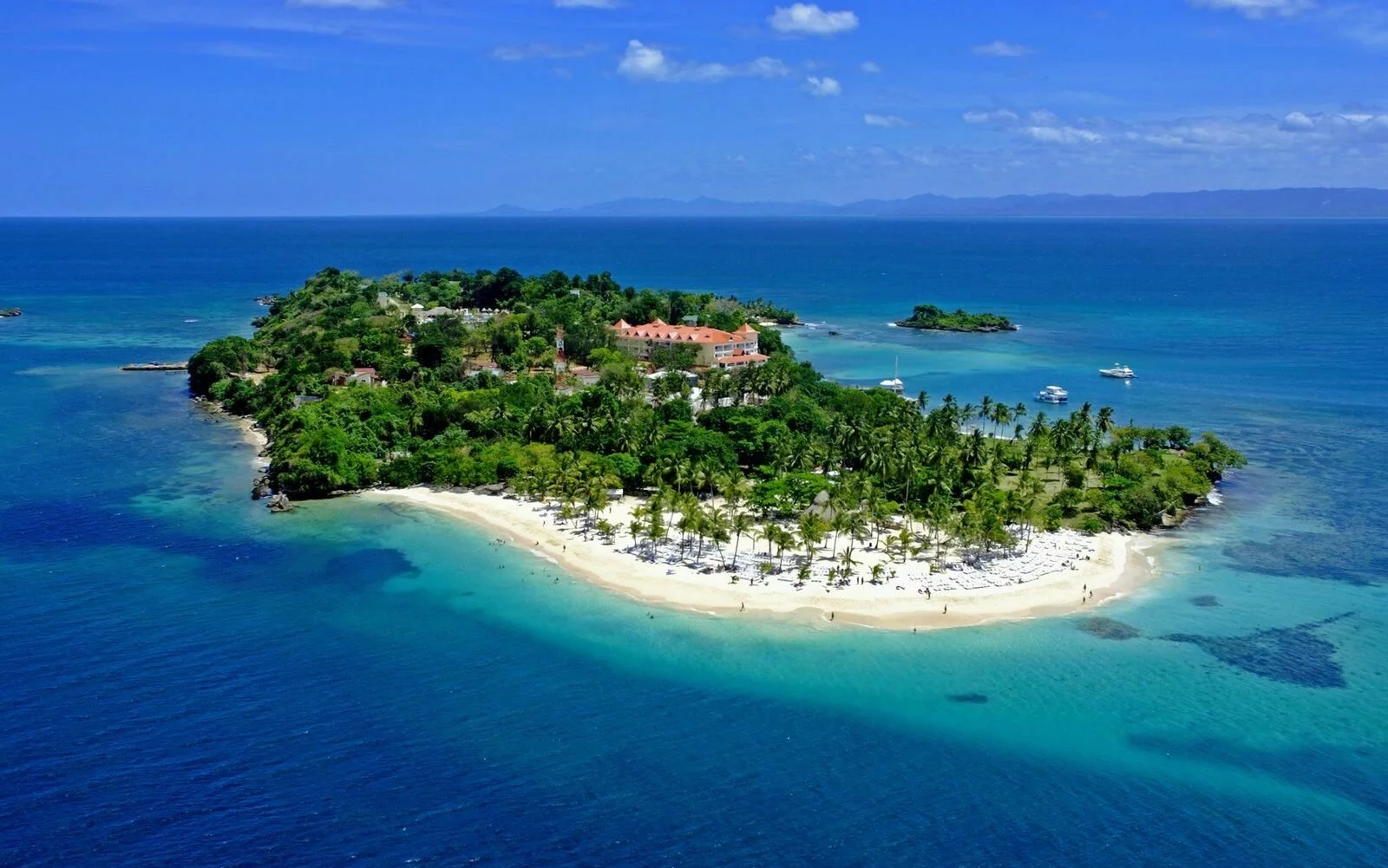 Aerial view of a tropical island with white sandy beaches, palm trees, and lush green vegetation, surrounded by clear blue ocean waters and boats anchored offshore.
