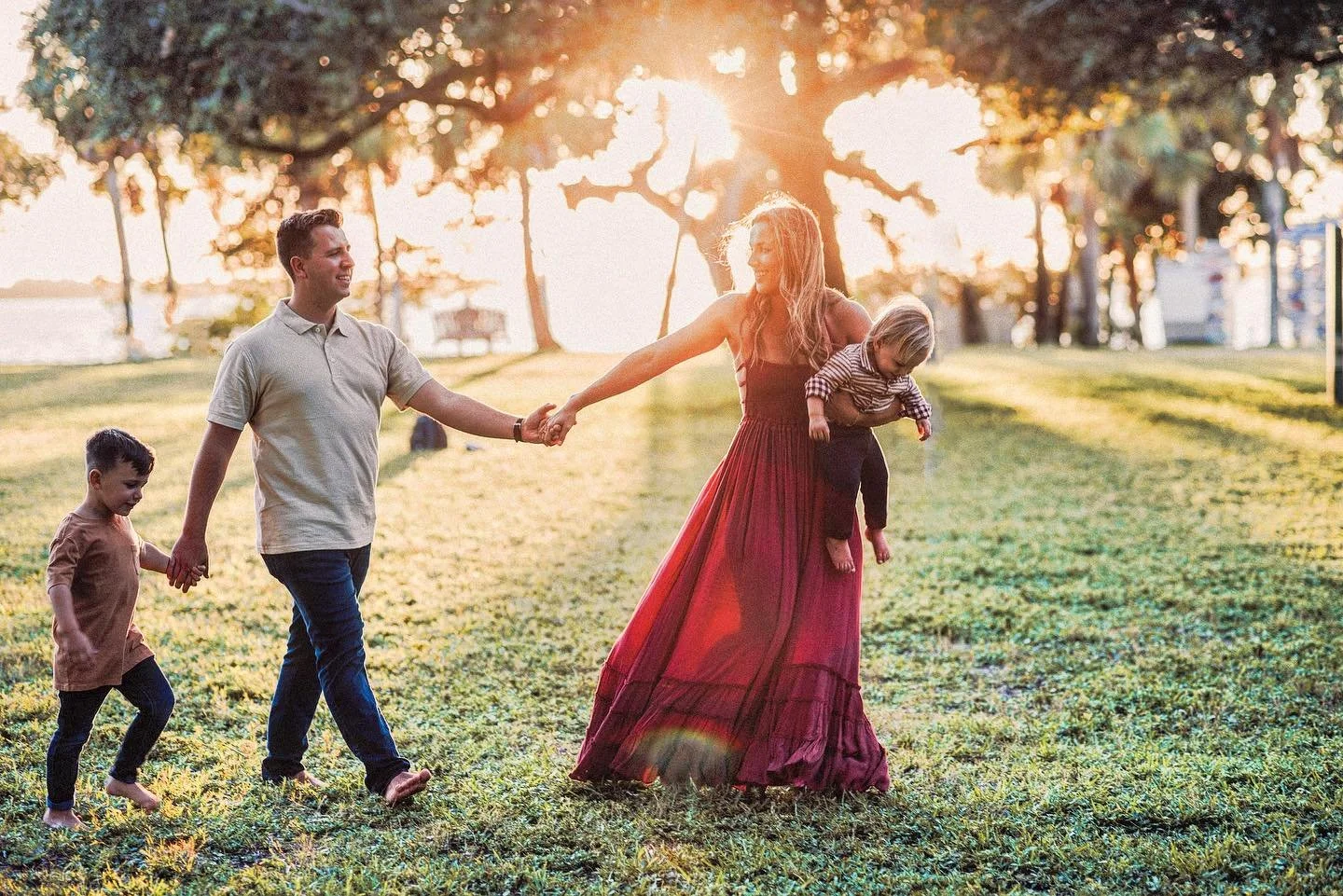A family of four enjoying a walk in a park during sunset, with a mother holding a child and holding hands with a father, and a young boy walking alongside them.