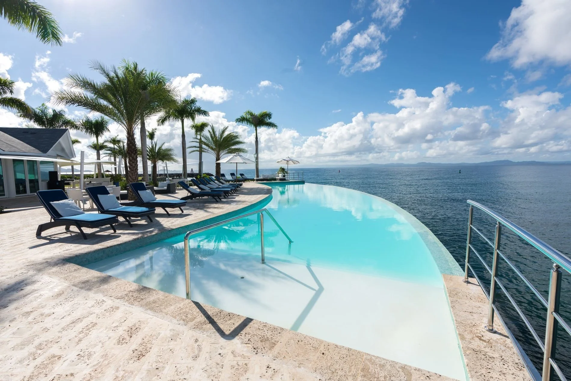 Infinity pool overlooking the ocean with lounge chairs and palm trees on a sunny day.