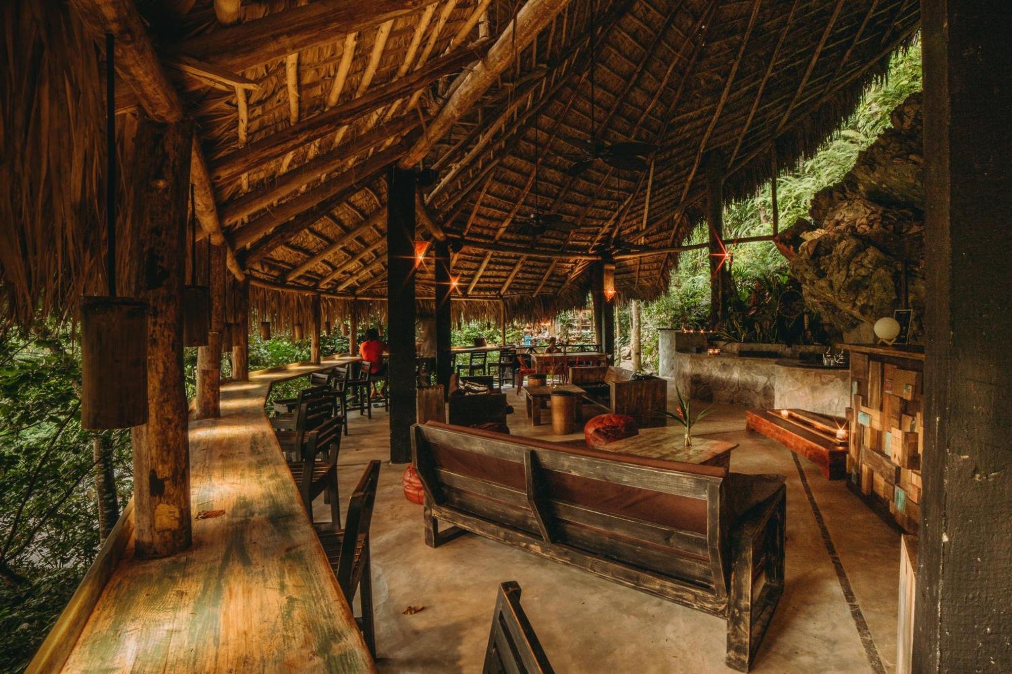 Interior of a rustic open-air bar or restaurant with wooden furniture, thatched roof, and lush greenery outside.