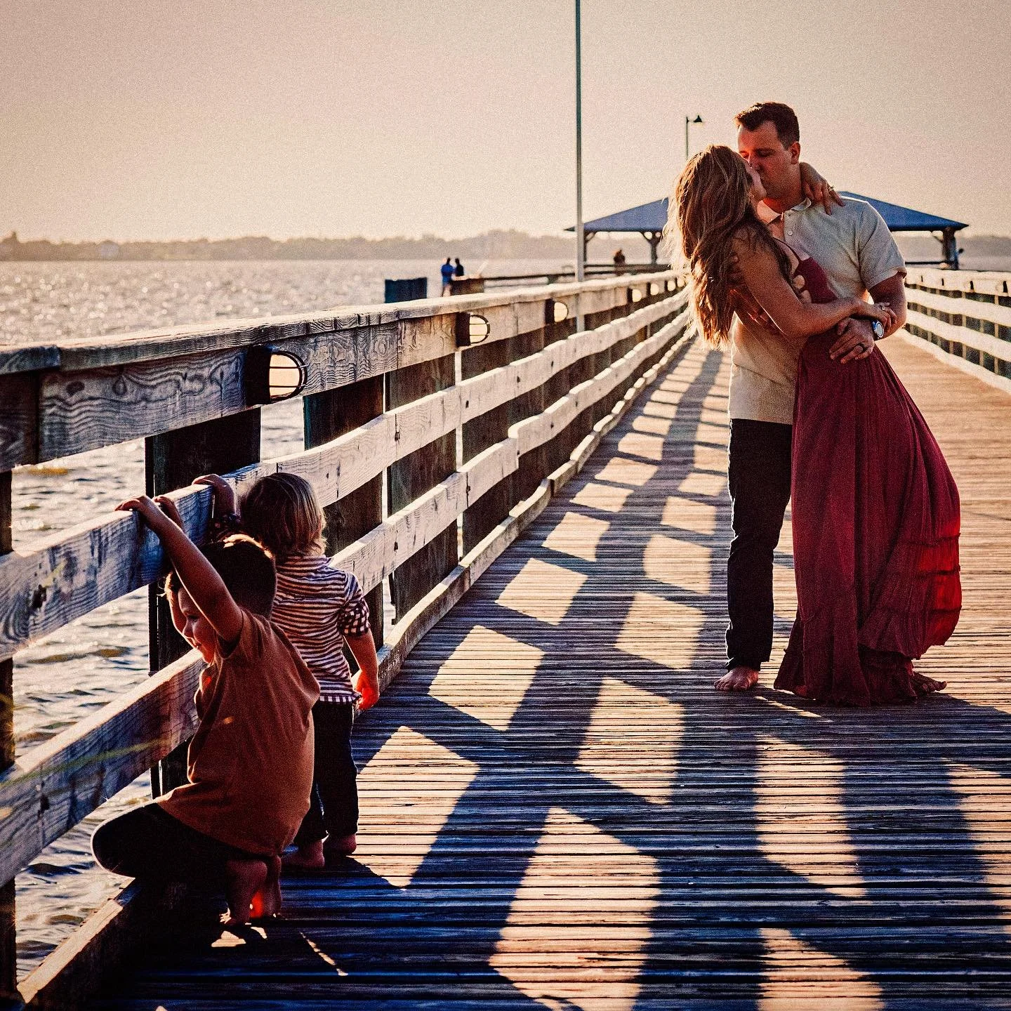 A couple kissing on a wooden pier at sunset with children playing nearby.
