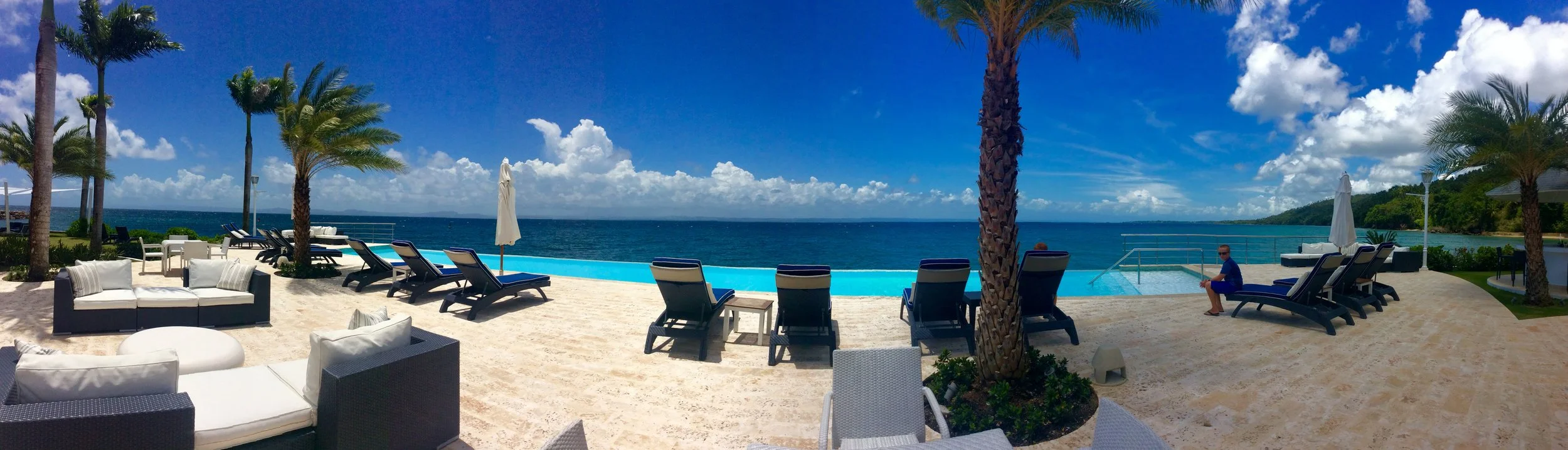 A beachside pool area with lounge chairs, umbrellas, and palm trees overlooking the ocean on a sunny day.