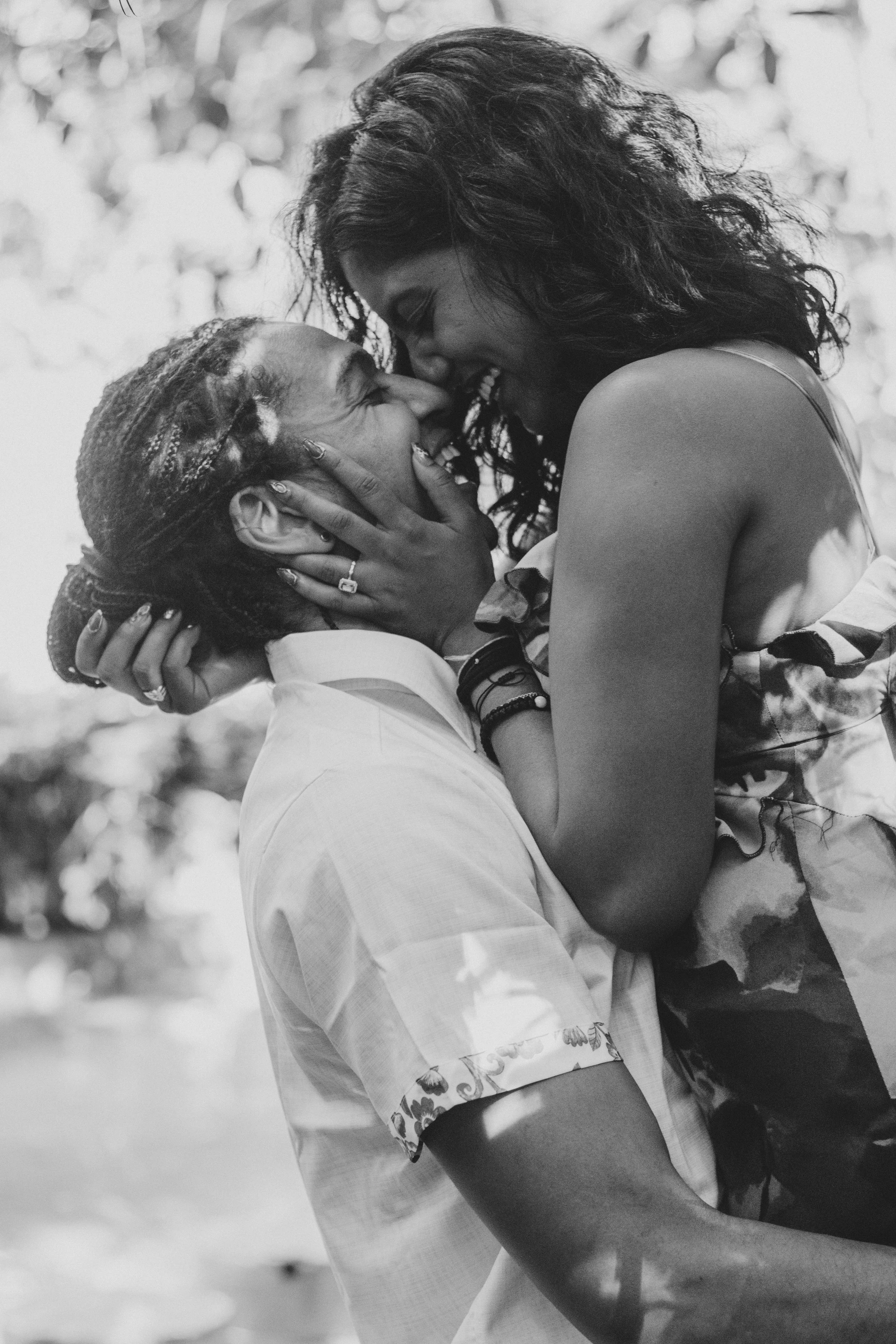 Black and white photo of a couple smiling and touching noses, the woman holding the man's face, outdoors with blurred trees in the background.
