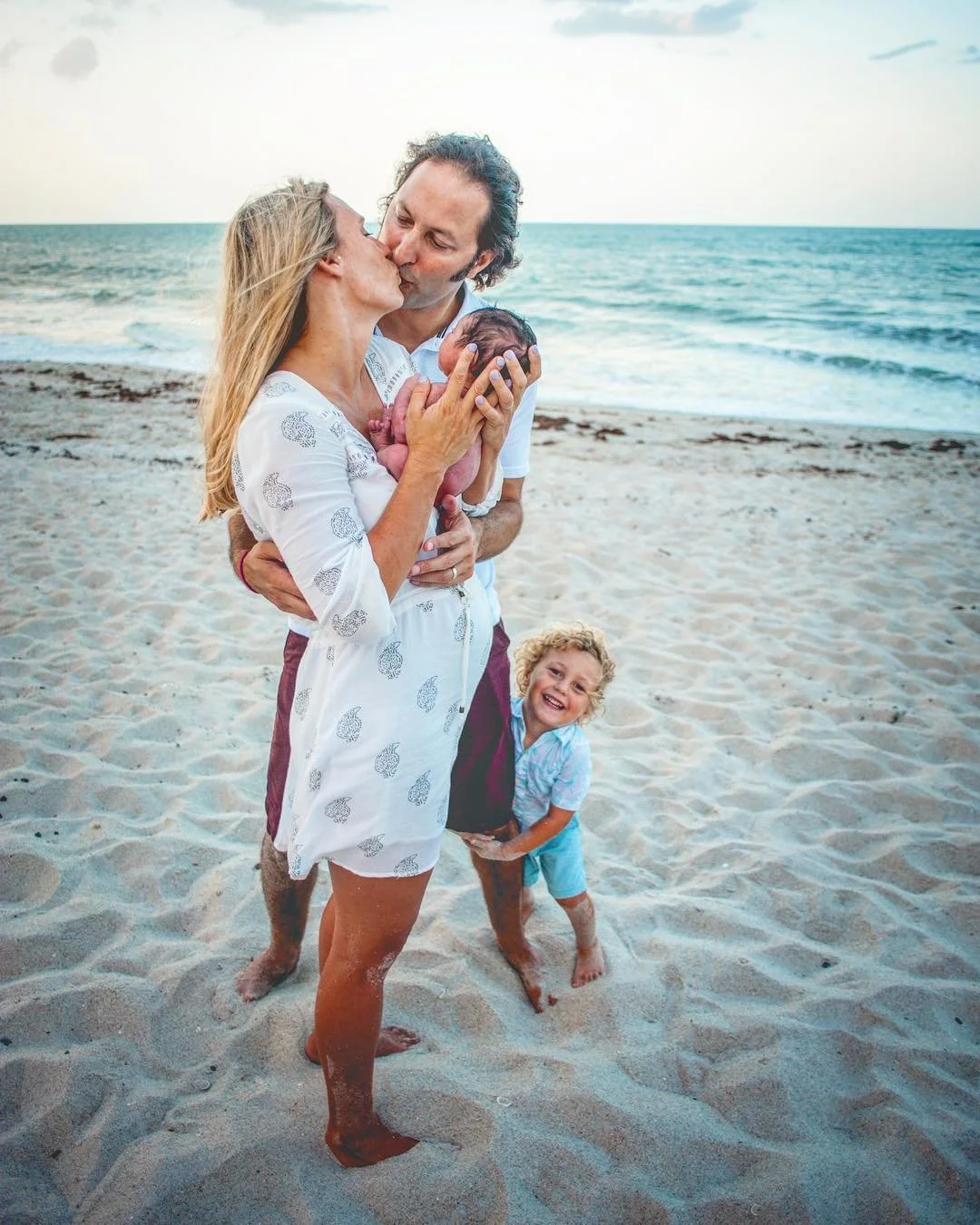A family on a beach: parents kissing while holding their newborn baby, with a smiling young boy standing nearby. The ocean and sky are in the background.