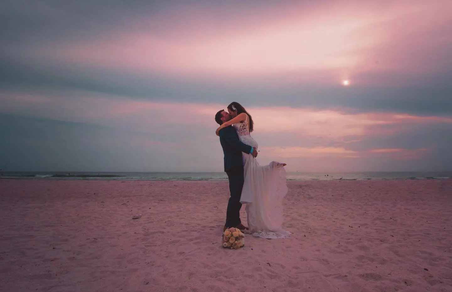 A couple in wedding attire embracing on a beach at sunset with a bouquet on the sand nearby.