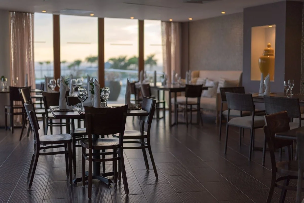 Indoor dining area with tables set with glasses and napkins, large windows with a view of trees, and modern decor with a neutral color palette.