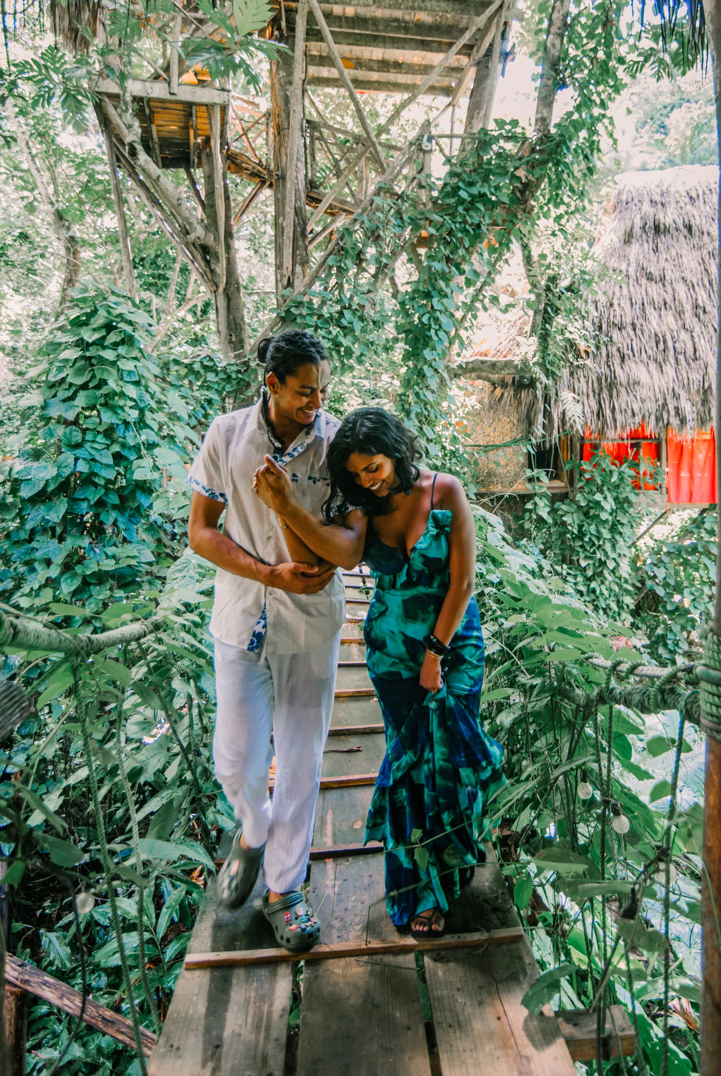 Two women walking on a wooden bridge surrounded by lush green foliage and trees in a jungle-like setting, smiling and enjoying each other's company.