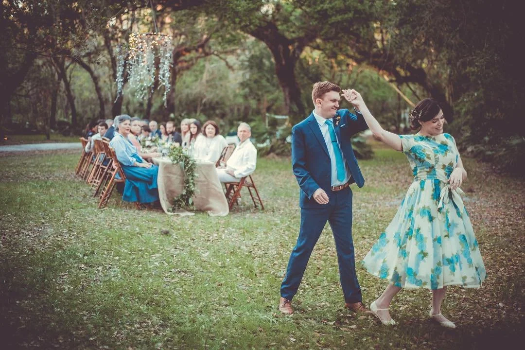 A couple dancing outdoors at a wedding reception with guests seated at a table in the background under trees.