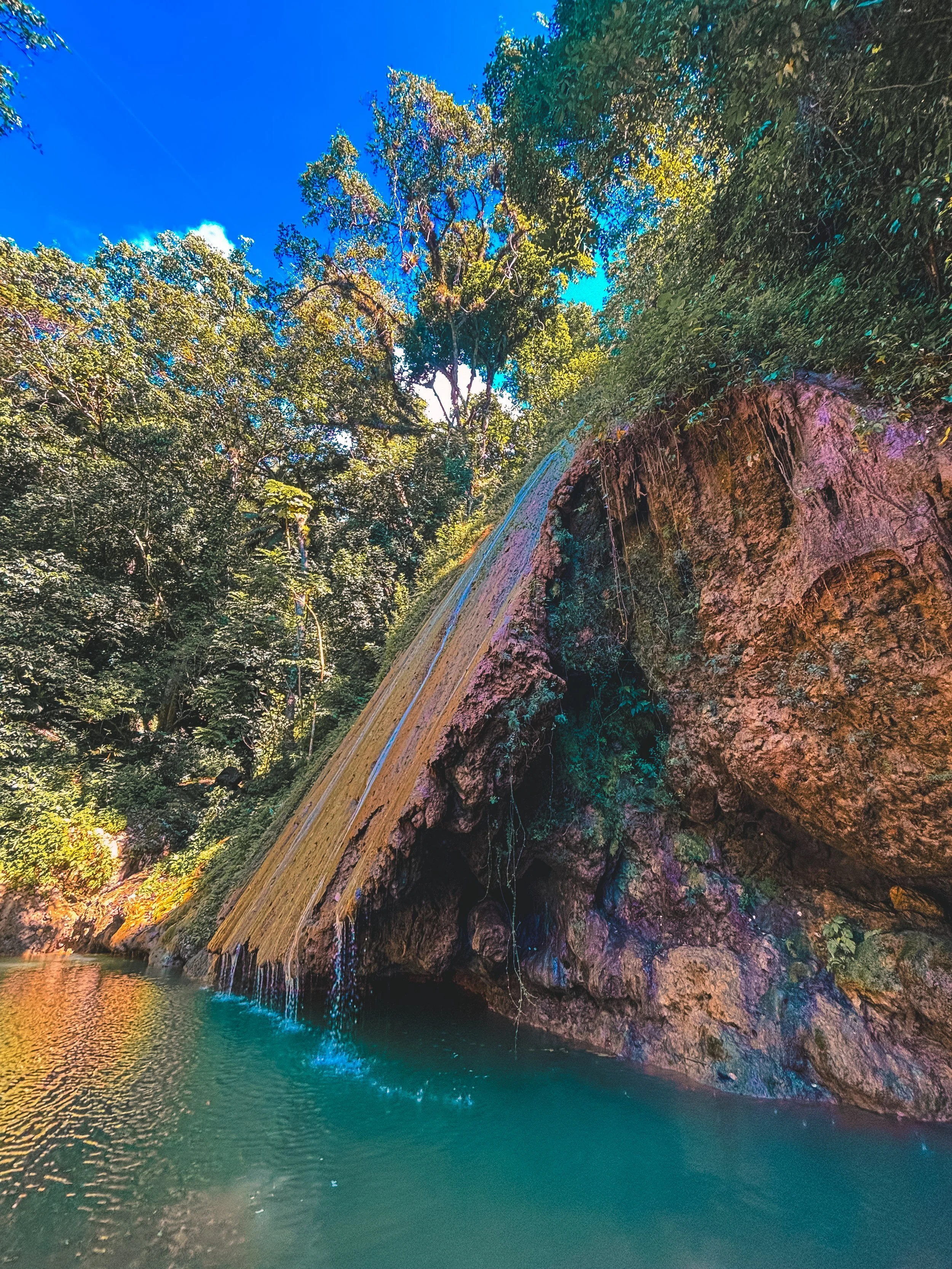 A small waterfall flows down a rocky cliff into a turquoise pool surrounded by lush green trees under a bright blue sky.