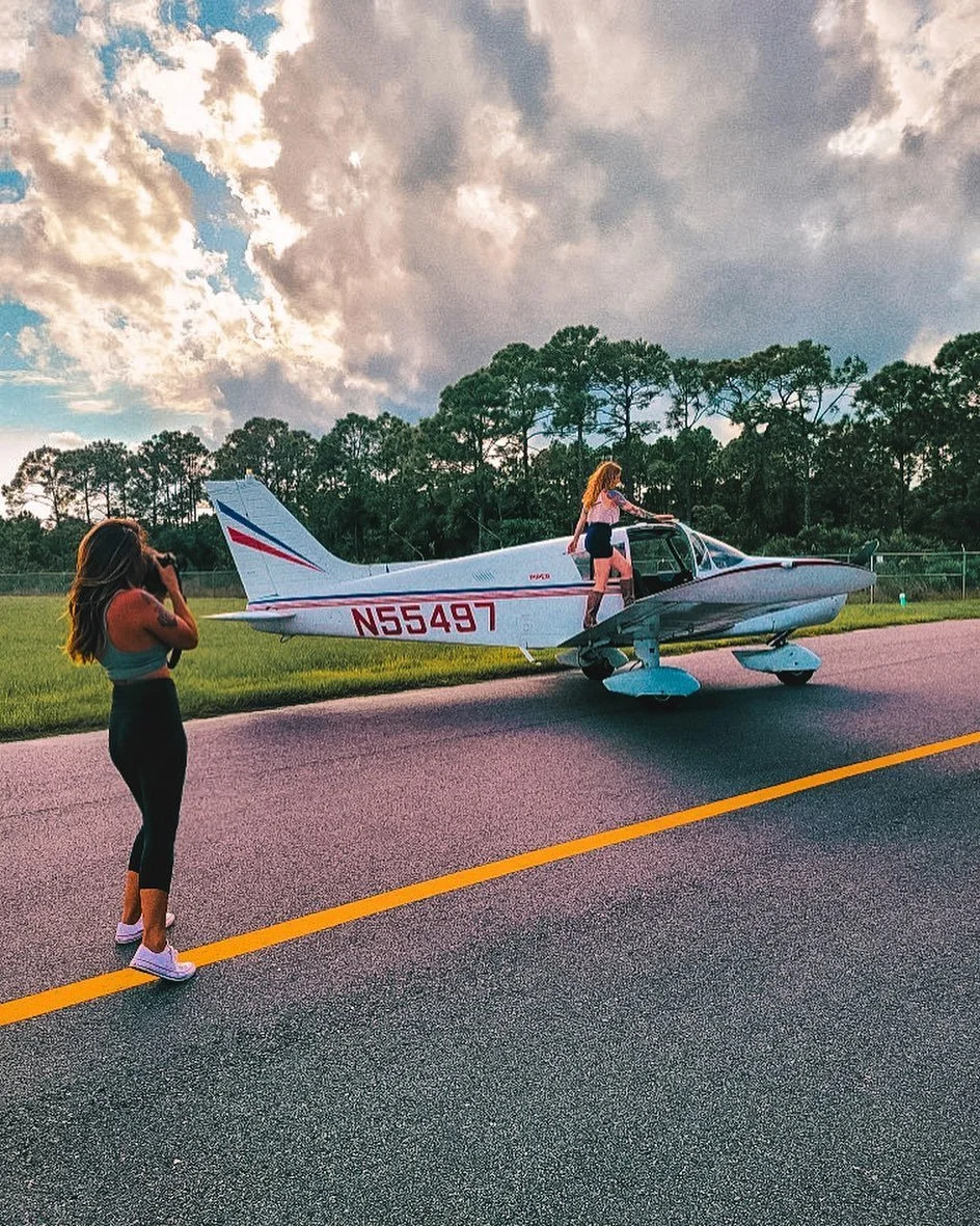 Two women near a small white and gray airplane, one standing on the ground and one climbing into the cockpit, on a paved runway during cloudy weather with tall trees in the background.