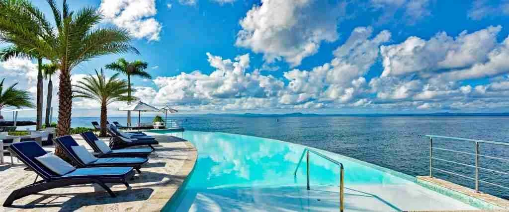 Poolside with lounge chairs and palm trees overlooking the ocean under a cloudy sky.