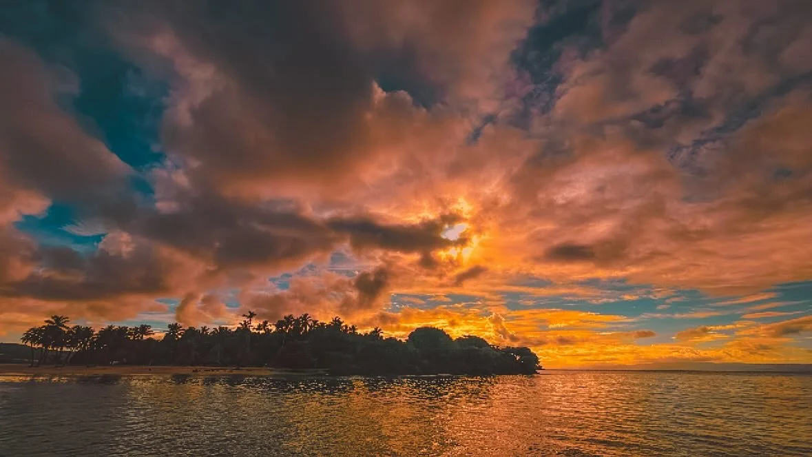 Sunset over a body of water with a silhouetted tree-lined shoreline and a colorful sky with clouds.
