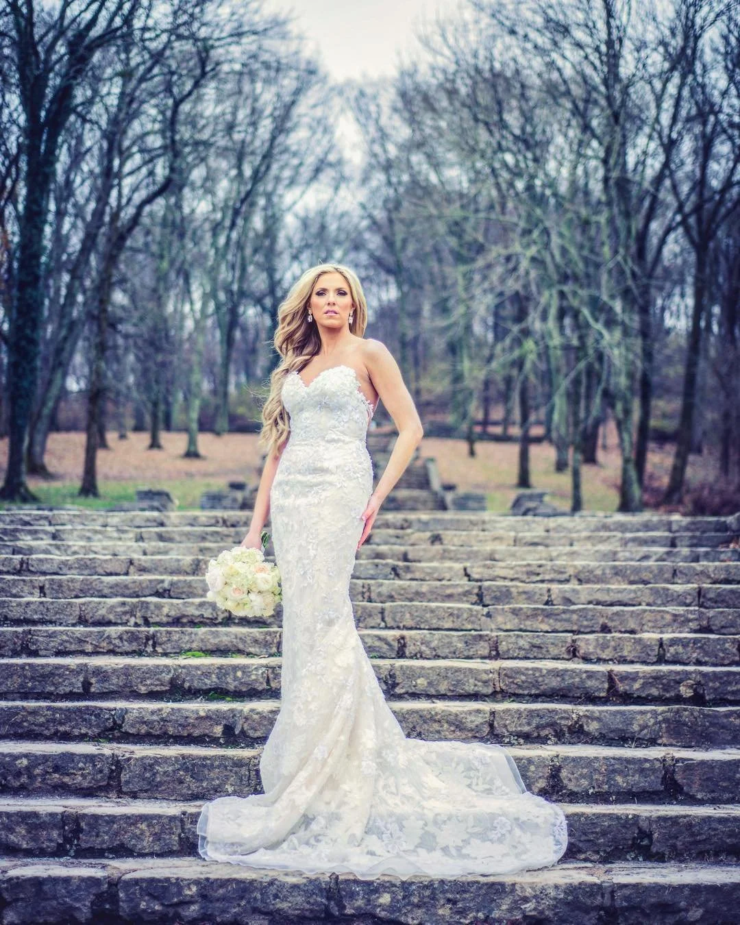 A bride in a white wedding dress holding a bouquet of white flowers stands on stone steps in an outdoor setting with leafless trees in the background.