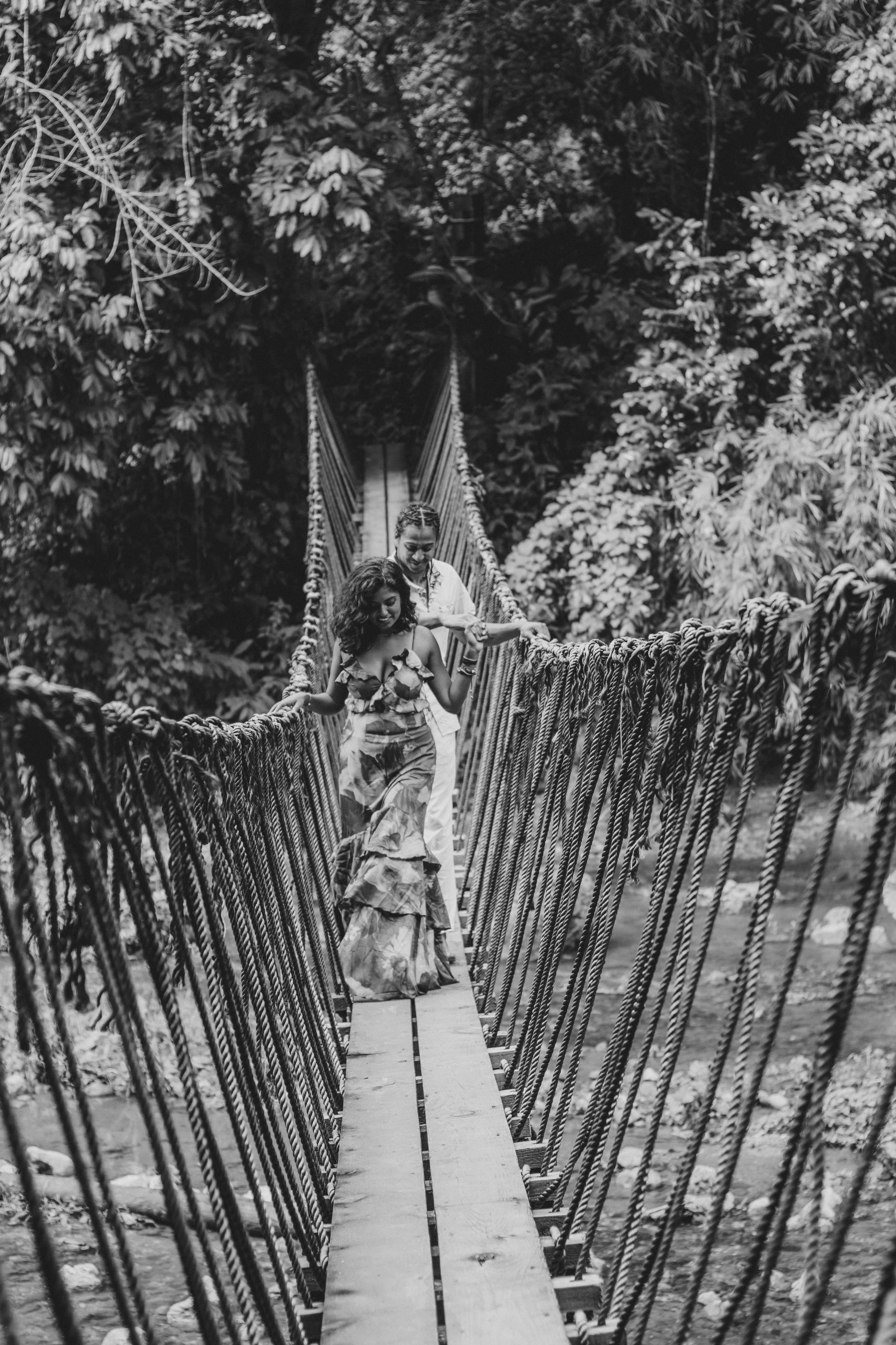 Two women walking across a hanging rope bridge in a lush, leafy jungle.