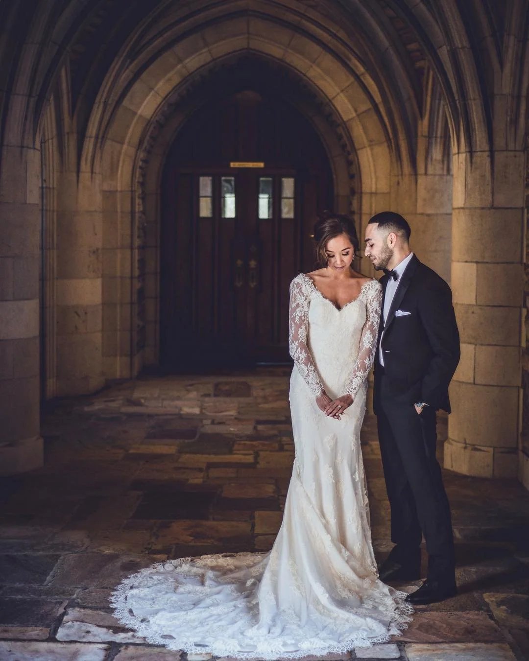 A bride in a lace wedding dress and a groom in a tuxedo standing close together, looking down, in front of a historic stone archway.