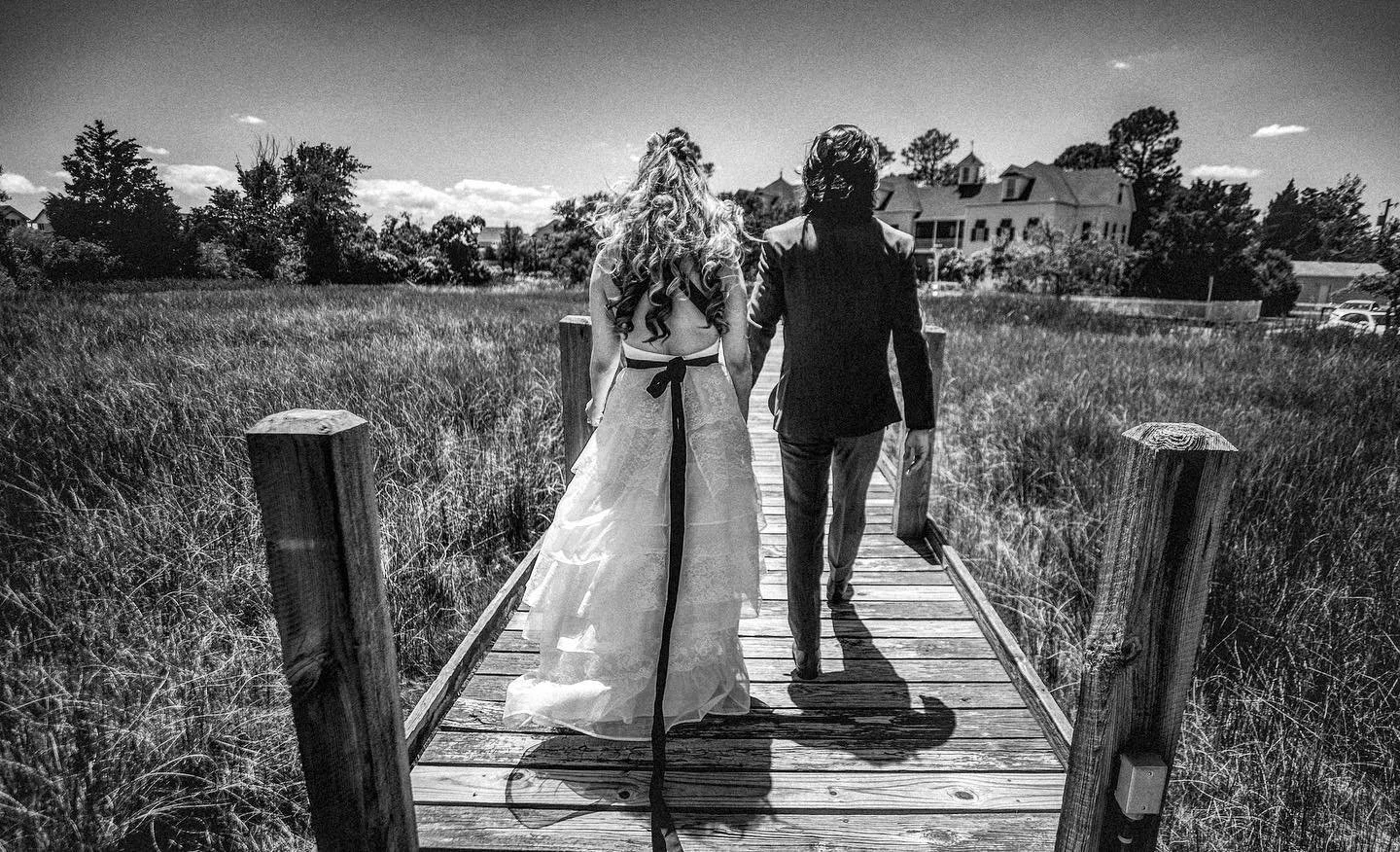 A black and white photo of a couple walking on a wooden bridge in a grassy field, with houses in the background.