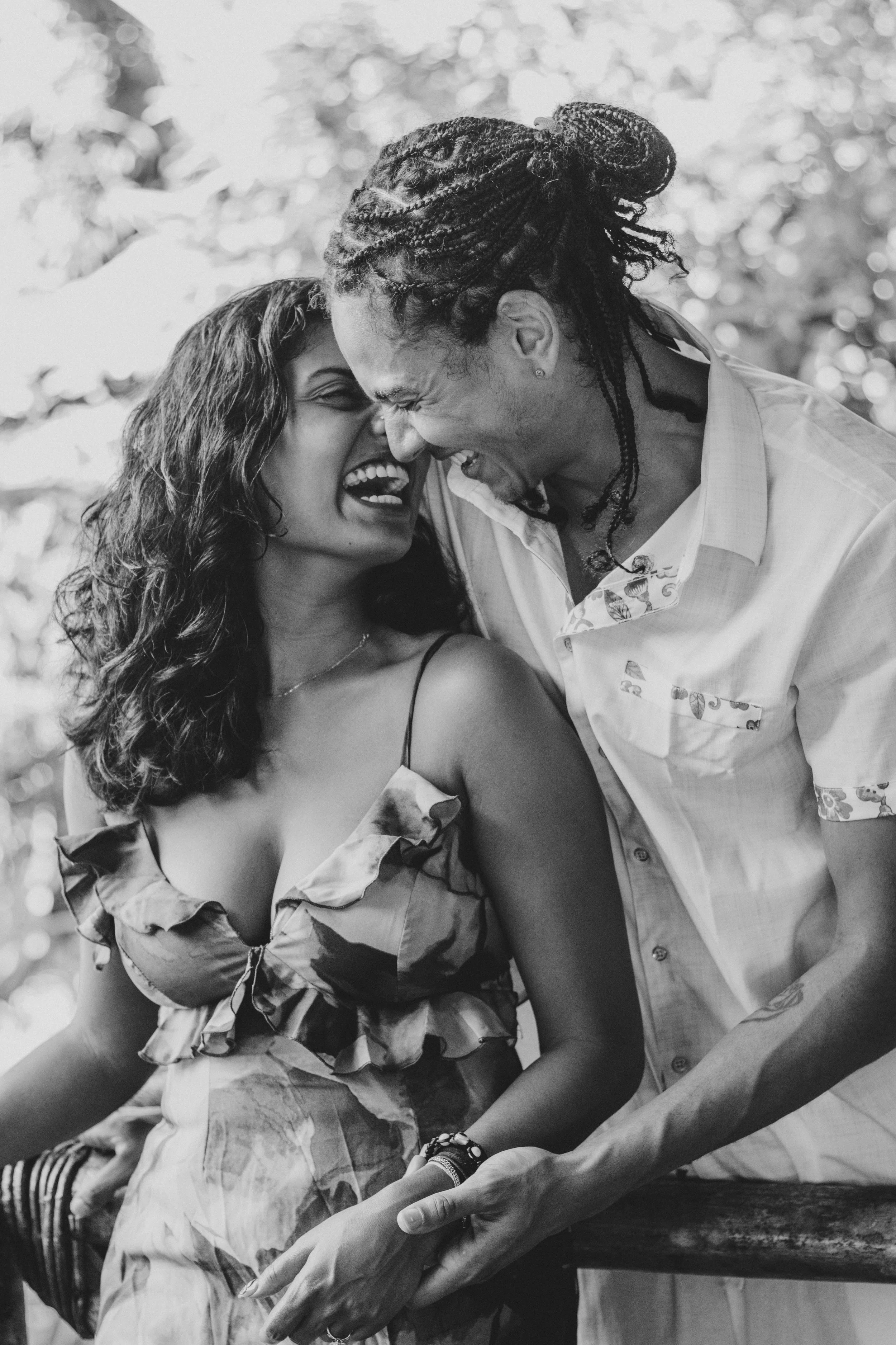 A black and white photo of a happy couple laughing together outdoors. The woman has curly hair and wears a ruffled dress, and the man with dreadlocks is leaning in close to her, both sharing a joyful moment.