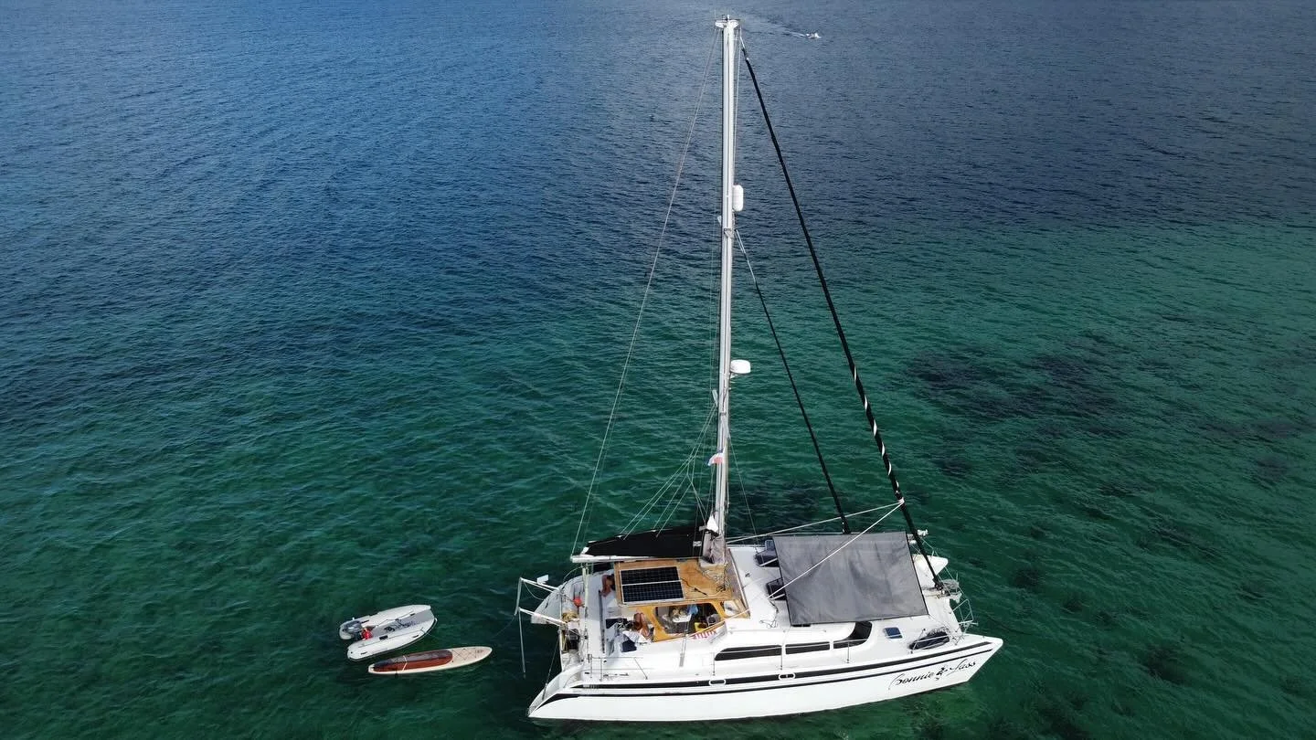 An aerial view of a white sailboat with a solar panel on the deck, floating on clear, green water near two smaller boats or kayaks attached to it.