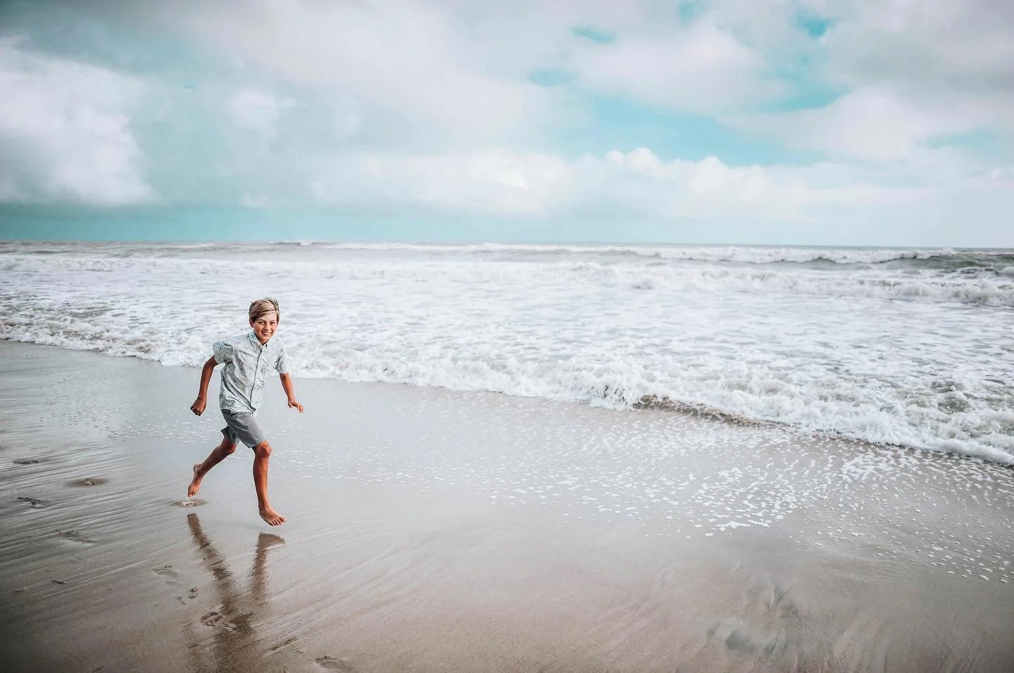 A young boy running barefoot along the wet sand of a beach near the ocean with waves crashing nearby and cloudy sky above.