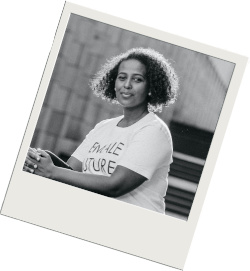 Eine Frau mit lockigem Haar, die in die Kamera lächelt, eine weiße T-Shirt trägt mit der Aufschrift 'FEMALE FUTURE', im Hintergrund eine Holzwand.