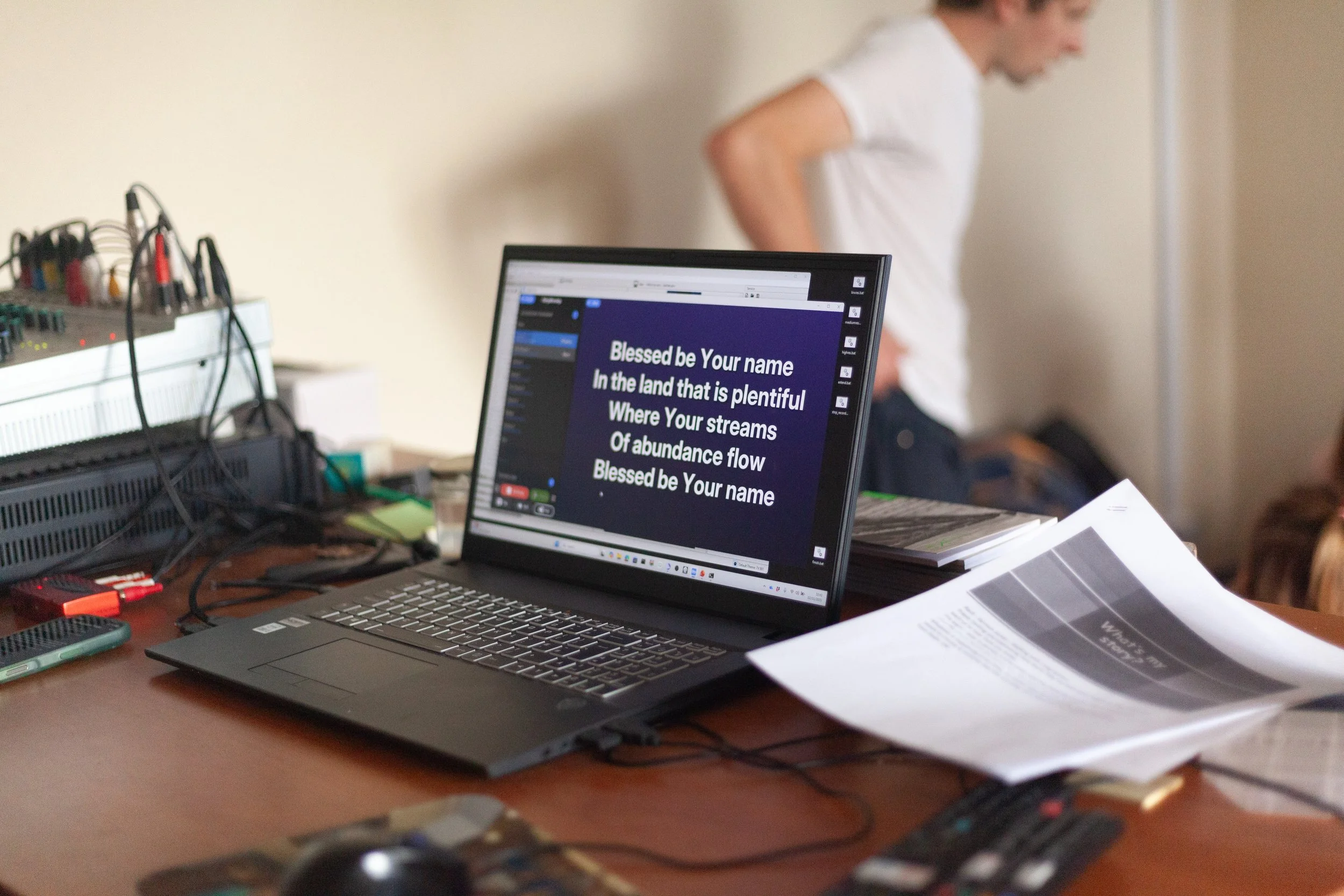 A laptop on a cluttered desk shows song lyrics on the screen, with a blurred person in a white shirt in the background.